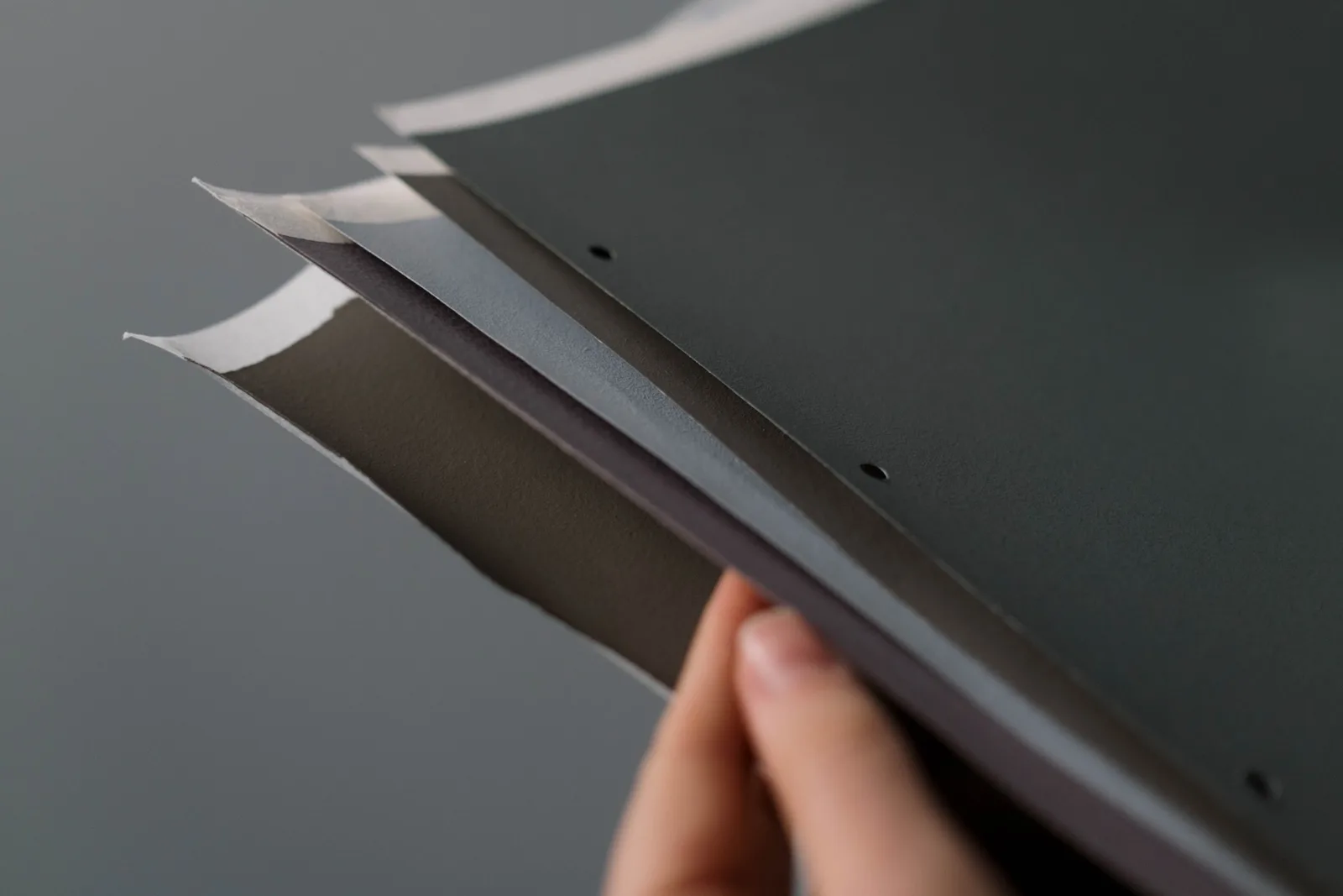 A close-up of a hand holding a set of gray paper samples against a neutral background. Book arts turns out to be four crafts sharing a single material