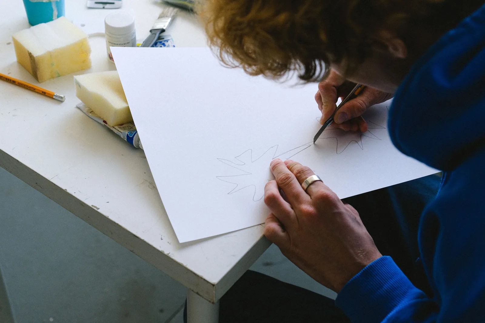 High angle of crop unrecognizable man in casual clothes sitting at table while cutting sketch on paper with utility knife near paints and sponge with pencil in light workshop. Paper marbling: four preparations that must line up before anything else